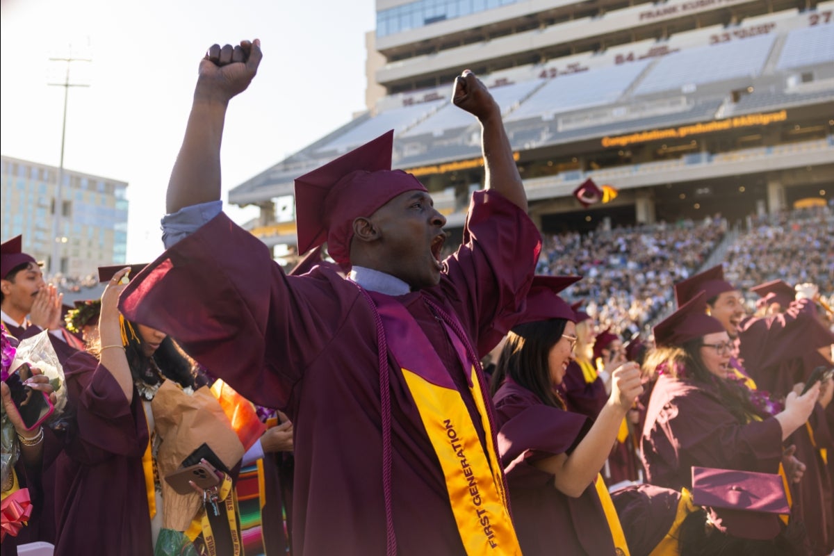 A man in graduation regalia raises his arms and cheers