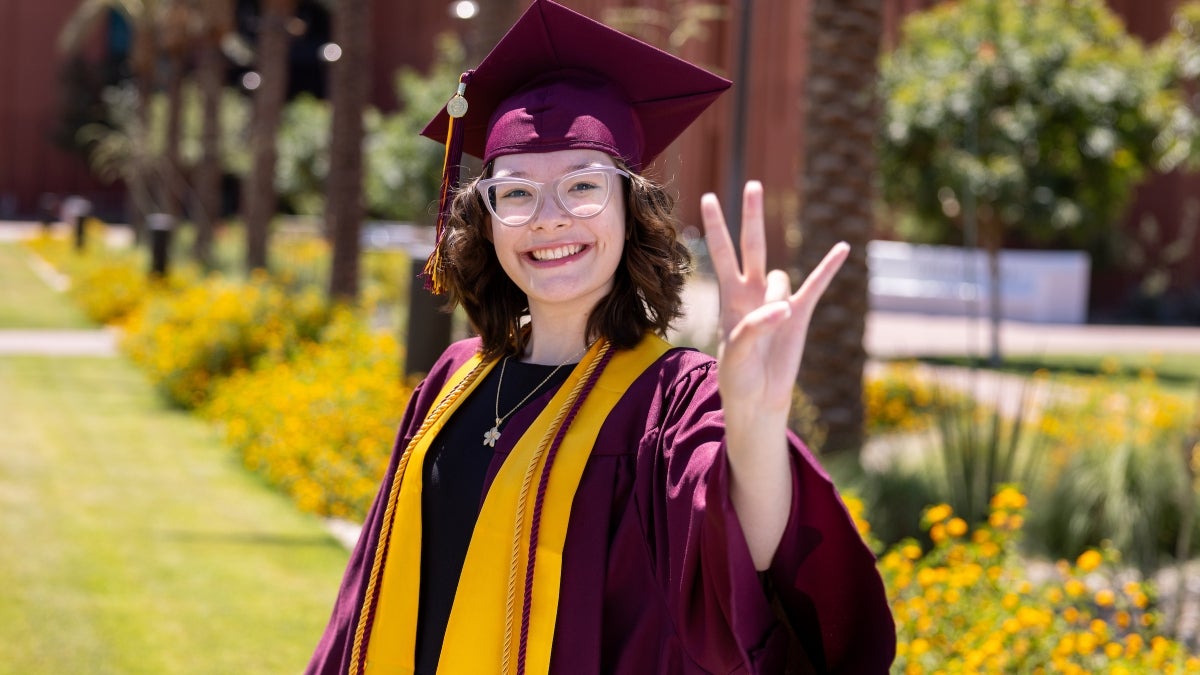Portrait of a young woman wearing a maroon cap and gown posing outdoors making a pitchfork sign with her hand