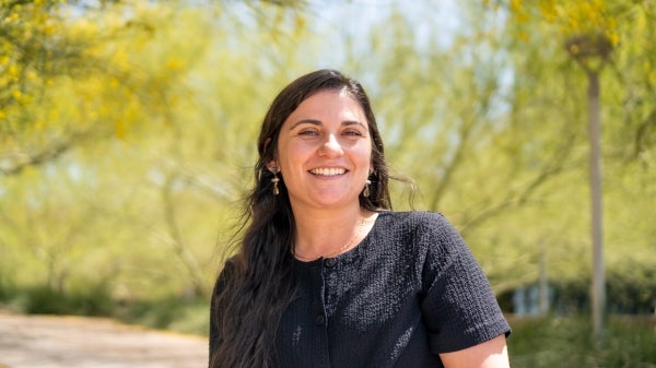 Holly Molinaro, wearing a black shirt and gold earrings, smiles in an outdoor setting with green leafy trees in the background.