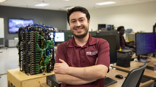 Ozgur Ozmen smiles with arms folded in a computer lab with equipment around him