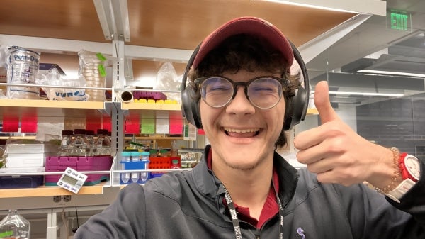 Alexander Sastokas, a Caucasian male student with brown hair, wearing headphones, a red ball cap, glasses and showing a thumbs up, standing in a laboratory in front of shelving with miscellaneous boxes