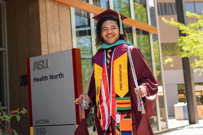 Dallas Salas smiles at the camera wearing his graduation cap and gown with all of his honor cords and stolls. He is sanding next to sign that says ASU Health North