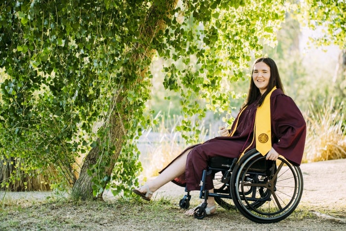 Kristen Bish wears her ASU graduation gown and stole. She is smiling at the camera while sitting in her wheelchair.